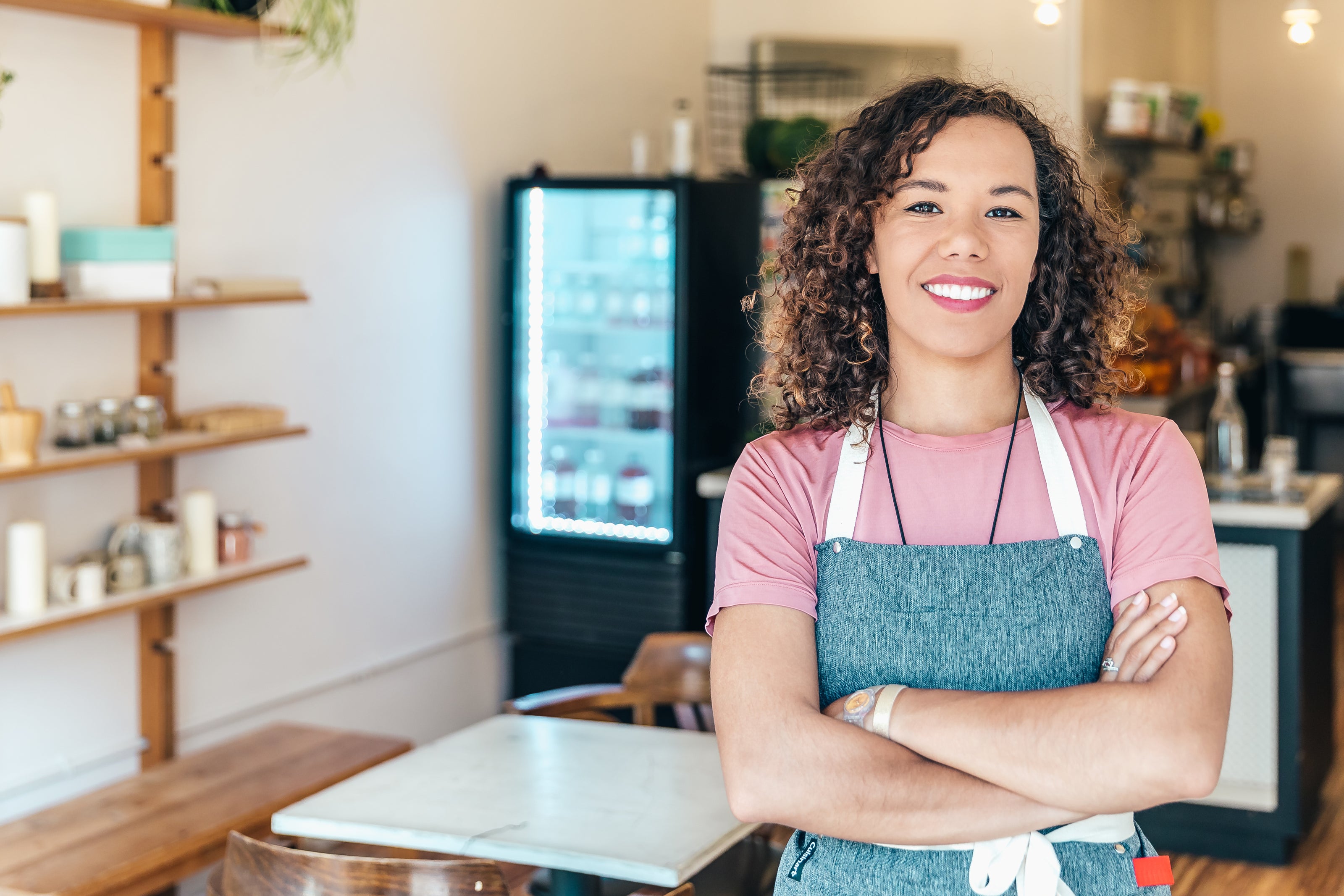 A smiling woman with curly hair wearing a gray apron and pink shirt stands confidently with her arms crossed inside a cozy café with wooden shelves and a refrigerator in the background.