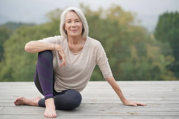 Middle-aged woman wearing comfortable clothing sits outdoors on a wooden deck in a relaxed yoga stretch pose with a calm expression and natural background of trees.