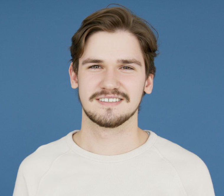 A young man with light brown hair and a short beard smiling gently, wearing a cream-colored sweater against a solid blue background.